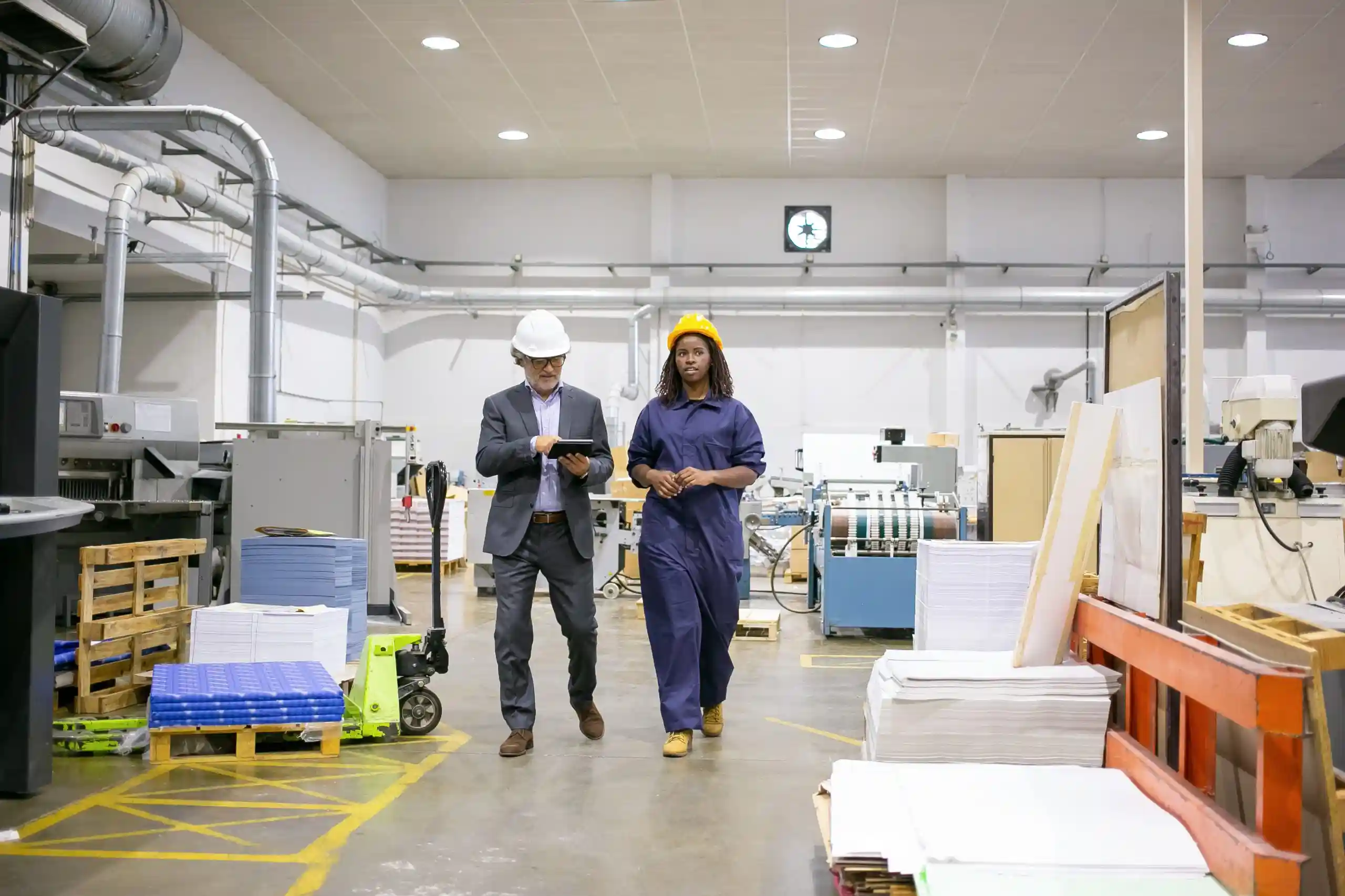 serious-male-inspector-female-factory-employee-hardhats-walking-plant-floor-talking-man-using-tablet-scaled